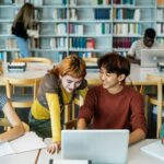 Young university students using laptop and studying with books in library - School education concept