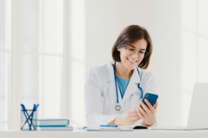 Positive female medical worker involved in chatting, holds mobile phone