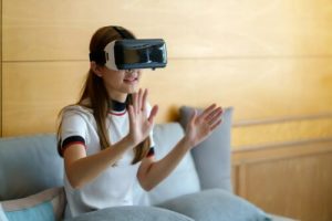 Smiling young Asian woman using VR glasses sitting on sofa at home.